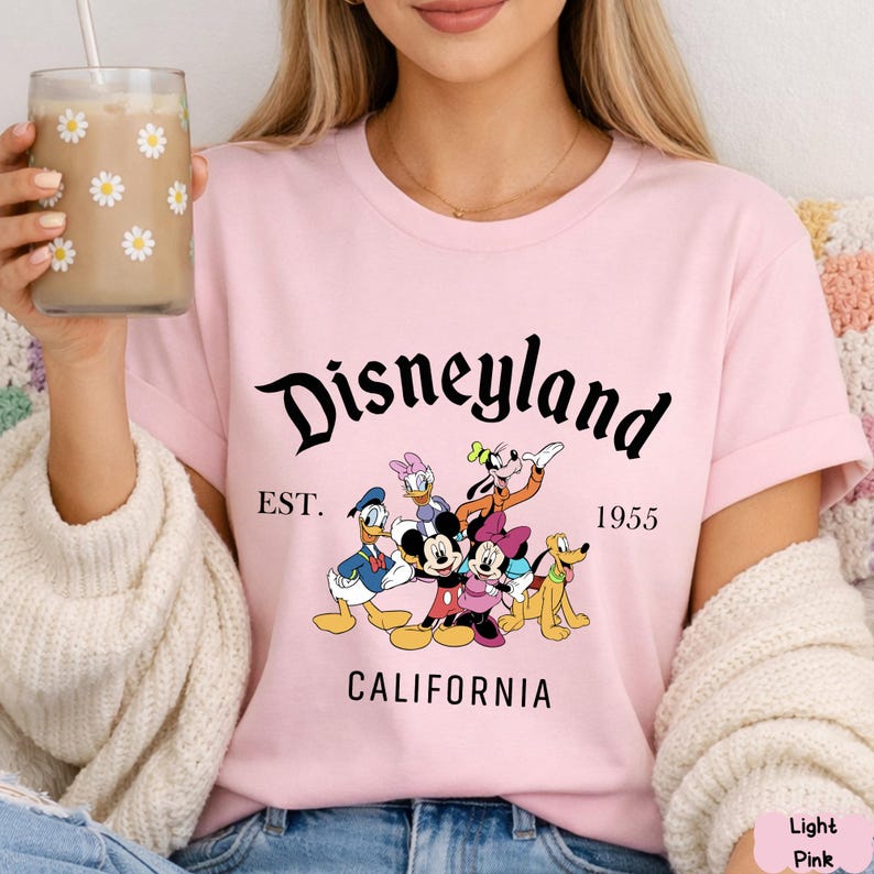May include: Light pink t-shirt featuring the word "Disneyland" in a black script font, with the text "EST. 1955 CALIFORNIA" below. The shirt displays a graphic of Disney characters including Mickey and Minnie Mouse, Donald Duck, and Goofy.