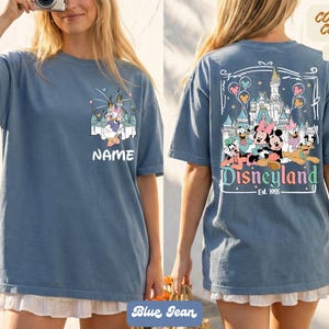 Puede incluir: Camiseta azul vaquero con dise&ntilde;o de Disneyland. La parte delantera tiene un gr&aacute;fico de dibujos animados con el nombre "NAME". La parte trasera presenta un castillo y personajes de Disneyland con el texto "Disneyland Est. 1955". Disponible en 19 colores.