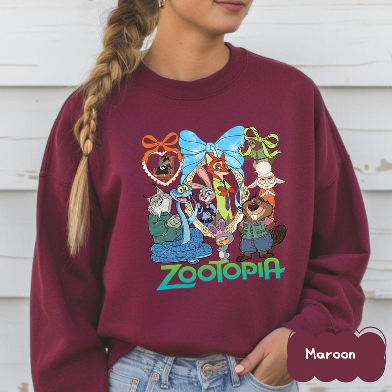 K&ouml;nnte beinhalten: Burgunderfarbenes Sweatshirt mit einer farbenfrohen Grafik von Charakteren aus dem Animationsfilm "Zoomania". Das Design umfasst eine gro&szlig;e blaue Schleife, das Wort "Zootopia" in Gr&uuml;n und verschiedene Tierfiguren. Das Sweatshirt ist einfarbig burgunderrot.