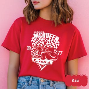 May include: A red t-shirt with a white graphic of Lightning McQueen from the Cars film. The design includes a race car, chequered flags, and the text "Lightning McQueen Cars 19 77".