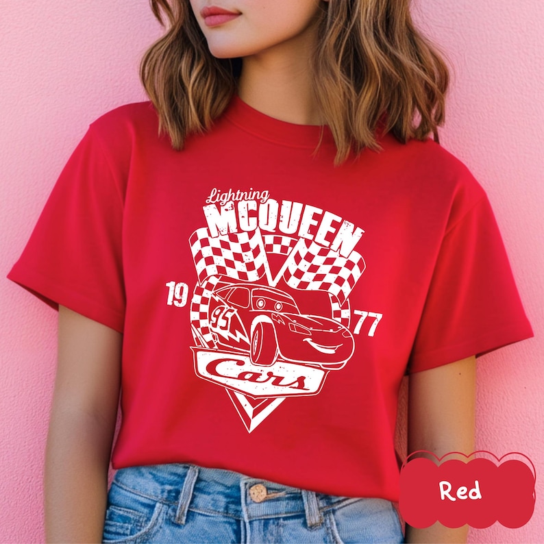 May include: A red t-shirt with a white graphic of Lightning McQueen from the Cars film. The design includes a race car, chequered flags, and the text "Lightning McQueen Cars 19 77".