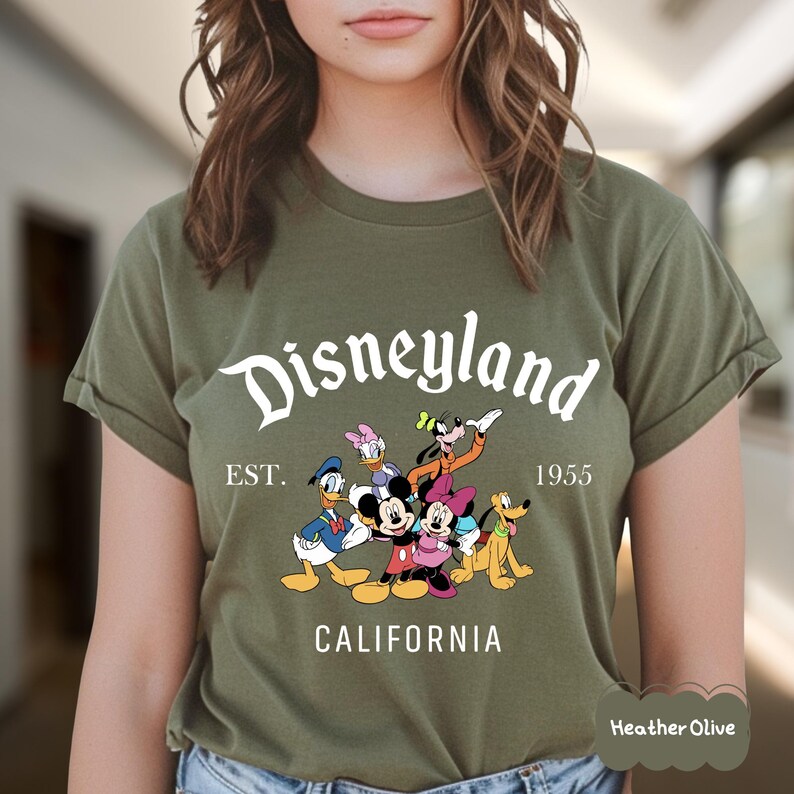 May include: Heather olive green t-shirt featuring the word "Disneyland" in a white script font, with the text "EST. 1955 CALIFORNIA" below. The shirt displays cartoon characters Mickey Mouse, Minnie Mouse, Donald Duck, Daisy Duck, Goofy, and Pluto.