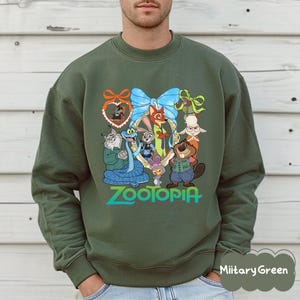 K&ouml;nnte beinhalten: Ein milit&auml;rgr&uuml;nes Sweatshirt mit einer farbenfrohen Grafik von Charakteren aus dem Animationsfilm "Zoomania". Das Design umfasst Judy Hopps, Nick Wilde und andere Charaktere, mit dem Wort "Zootopia" in Gr&uuml;n.