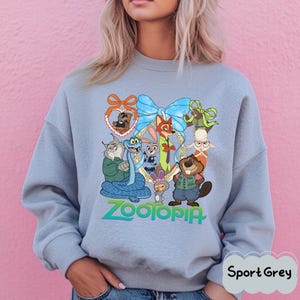 K&ouml;nnte beinhalten: Sportgraues Sweatshirt mit einem farbenfrohen Aufdruck von Charakteren aus dem Film Zootopia. Das Design umfasst eine blaue Schleife, einen goldenen Rahmen und das Wort "Zootopia" in Gr&uuml;n. Das Sweatshirt ist hellgrau.