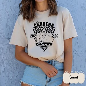 May include: Sand-coloured t-shirt with a graphic of Sally Carrera from the film Cars. The design includes the text "Sally Carrera," "Cars," and the numbers "2002" with a chequered flag background. The shirt has a crew neck.