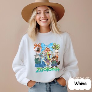 K&ouml;nnte beinhalten: Wei&szlig;es Sweatshirt mit einer farbenfrohen Grafik von Charakteren aus dem Animationsfilm "Zoomania". Das Design umfasst eine blaue Schleife, verschiedene Tierfiguren und das Wort "Zootopia" in Gr&uuml;n. Das Sweatshirt hat einen Rundhalsausschnitt.