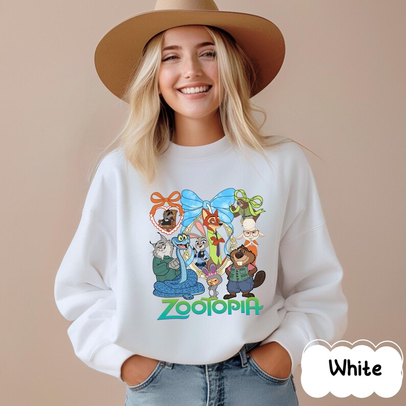 K&ouml;nnte beinhalten: Wei&szlig;es Sweatshirt mit einer farbenfrohen Grafik von Charakteren aus dem Animationsfilm "Zoomania". Das Design umfasst eine blaue Schleife, verschiedene Tierfiguren und das Wort "Zootopia" in Gr&uuml;n. Das Sweatshirt hat einen Rundhalsausschnitt.