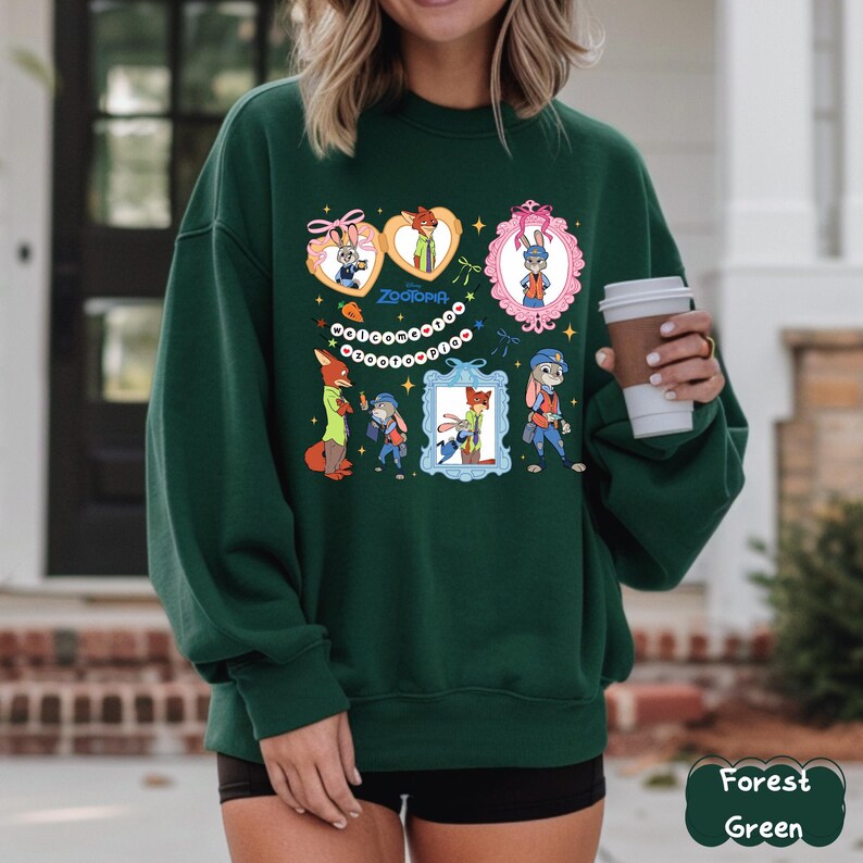 May include: Forest green sweatshirt featuring Zootopia characters in decorative frames and heart shapes. The design includes Nick Wilde, Judy Hopps, and other characters from the animated film. The word "Zootopia" is also printed on the sweatshirt.