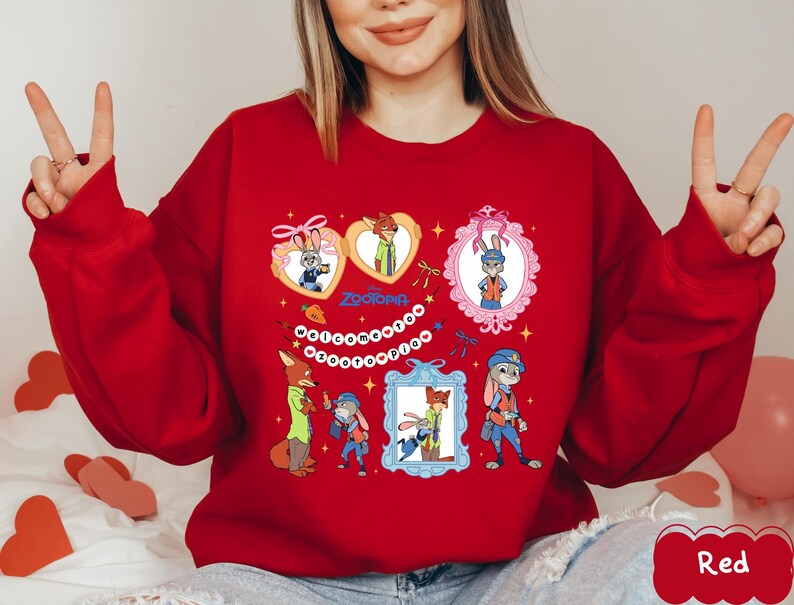 May include: A red sweatshirt featuring cartoon characters from the movie Zootopia. The design includes images of a fox and a rabbit in various frames and heart shapes, with the word "Zootopia" in the center. The sweatshirt has long sleeves.