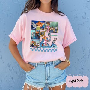 May include: Light pink t-shirt featuring a colorful graphic of characters from the animated movie Zootopia. The design includes multiple panels with images of a fox, a rabbit, and other characters. The word "Zootopia" is written in green.