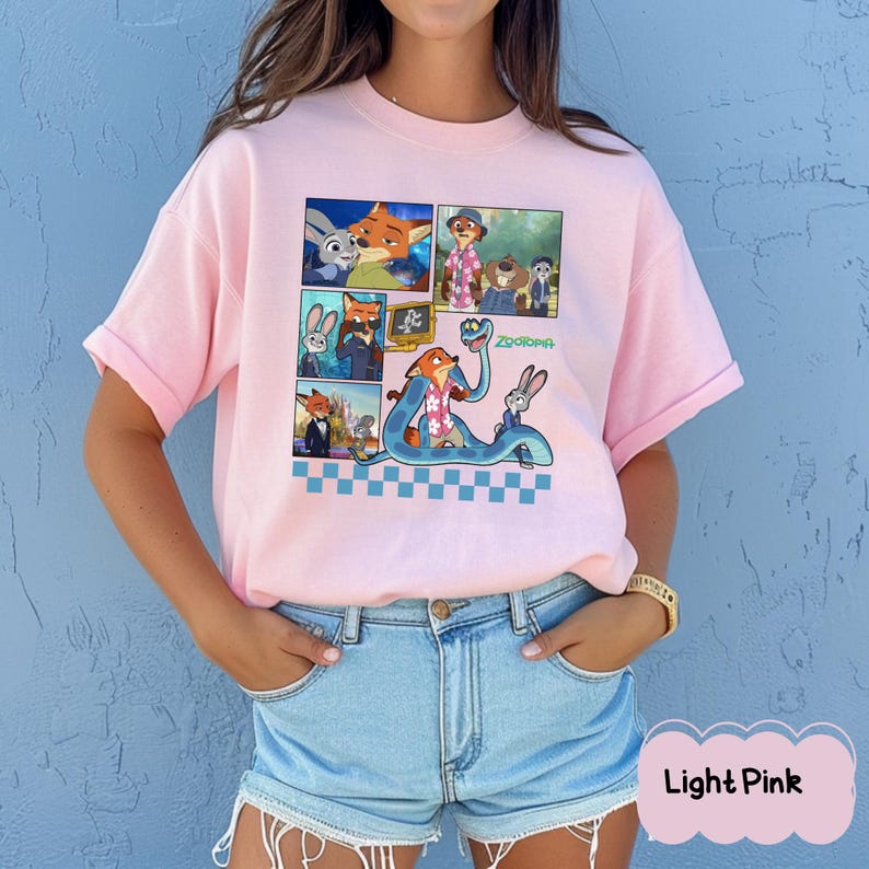May include: Light pink t-shirt featuring a colorful graphic of characters from the animated movie Zootopia. The design includes multiple panels with images of a fox, a rabbit, and other characters. The word "Zootopia" is written in green.