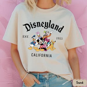 May include: Sand-colored t-shirt featuring the word "Disneyland" in a stylized font, with "EST. 1955" and "CALIFORNIA" below. The shirt displays a colorful graphic of Disney characters, including Mickey and Minnie Mouse, Donald Duck, and Goofy.