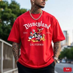 May include: A red t-shirt featuring the Disneyland logo and a graphic of Disney characters, including Mickey Mouse, Minnie Mouse, Donald Duck, and Goofy. The text "EST. 1955 CALIFORNIA" is also printed on the shirt.