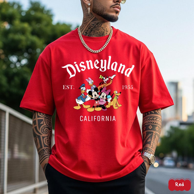 May include: A red t-shirt featuring the Disneyland logo and a graphic of Disney characters, including Mickey Mouse, Minnie Mouse, Donald Duck, and Goofy. The text "EST. 1955 CALIFORNIA" is also printed on the shirt.