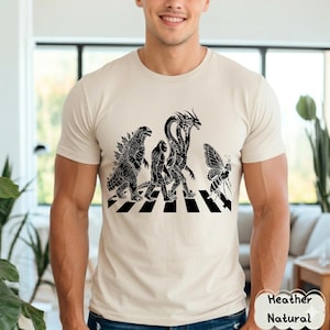 May include: Beige t-shirt featuring a black and white graphic of Godzilla, a Bigfoot-like creature, a three-headed dragon, and a butterfly crossing a crosswalk. The shirt is made of a soft, neutral-colored material.
