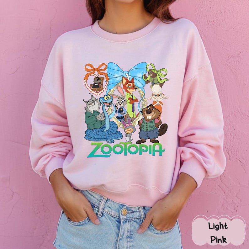 K&ouml;nnte beinhalten: Hellrosa Sweatshirt mit Rundhalsausschnitt und farbenfroher Grafik von Charakteren aus dem Film "Zootopia". Das Design beinhaltet eine gro&szlig;e blaue Schleife und das Wort "Zootopia" in gr&uuml;ner und blauer Schrift. Das Sweatshirt ist aus weichem Material.