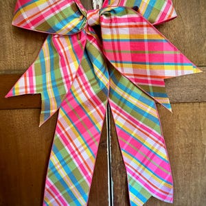 May include: A large decorative bow with a vibrant plaid pattern in pink, green, yellow, and blue. The bow is tied in the center with long, pointed tails. The bow is likely used for decorating, gift wrapping, or seasonal displays.