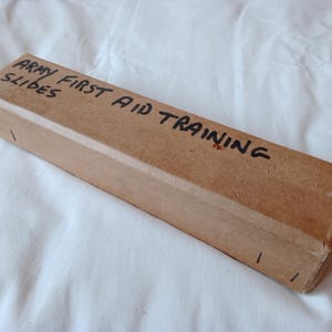 May include: A rectangular cardboard box with the text "ARMY FIRST AID TRAINING SLIDES" in black ink. The box is a light brown colour and appears to be closed. The box is resting on a white, wrinkled fabric.