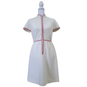 May include: A white knee-length dress with short sleeves and a high collar. The garment features a vertical red and white patterned trim running down the center front and around the waist, with matching trim on the sleeve edges.