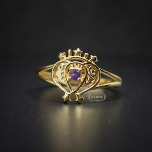 Puede incluir: Un anillo de oro con una piedra preciosa púrpura central. El anillo presenta un diseño ornamentado con detalles de estrellas y un filigrana en forma de corazón. El anillo está sobre un fondo oscuro, resaltando sus detalles.