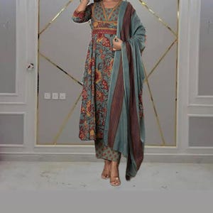 May include: A teal-colored, floral-patterned kurta suit with matching pants and a shawl. The kurta features a V-neck and three-quarter sleeves. The shawl has a burgundy and brown striped border. The outfit is suitable for festive occasions.