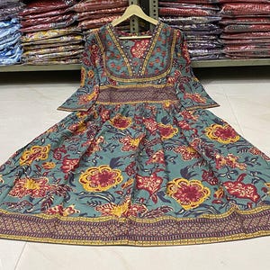May include: A teal, floral-print dress with a V-neck and long sleeves. The dress features a pattern of red and yellow flowers and a decorative border. The dress is hanging on a wooden hanger.