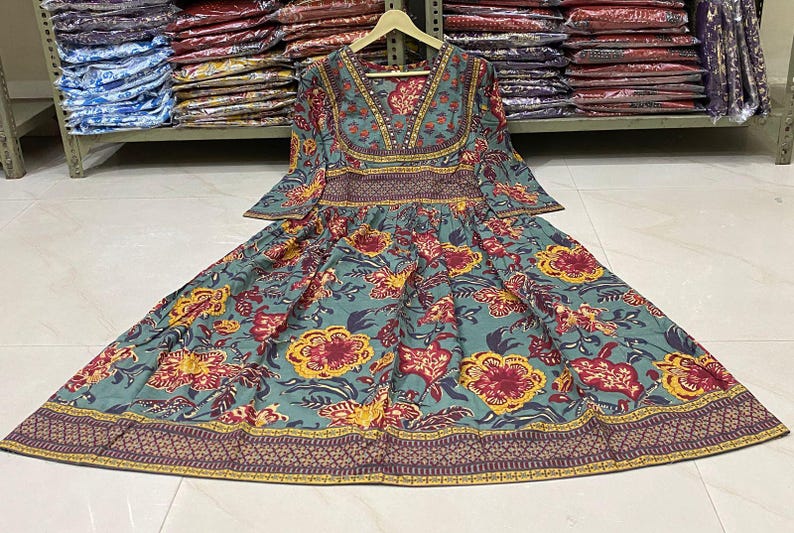 May include: A teal, floral-print dress with a V-neck and long sleeves. The dress features a pattern of red and yellow flowers and a decorative border. The dress is hanging on a wooden hanger.