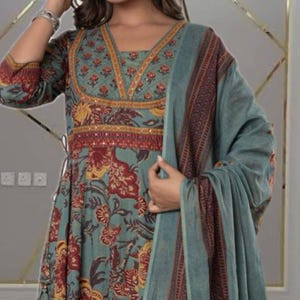 May include: A teal-colored, long-sleeved dress with a floral pattern in red and yellow. The dress features a V-neckline with intricate gold and red embroidery. A matching teal shawl with a patterned border drapes over the shoulders.