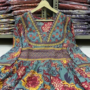 May include: A teal-colored long-sleeve dress with a V-neck and floral pattern. The dress features a mix of red, yellow, and teal floral designs, with decorative bands around the neckline and waist. The dress is hanging on a wooden hanger.