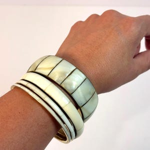 May include: A set of three stacked bangle bracelets featuring a wide band with a segmented mother-of-pearl inlay, a smooth off-white band, and a thin metallic band. The bracelets have a polished finish and are worn together on the wrist.