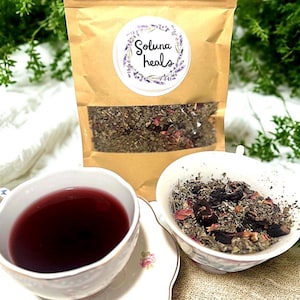 Könnte beinhalten: Ein brauner Papiersack mit einem transparenten Fenster, das getrocknete Kräuter und Blumen zeigt, neben einer Teetasse mit roter Flüssigkeit und einer kleinen Schüssel mit Kräutern. Der Beutel trägt die Aufschrift "Soluna heals".