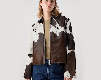 May include: A brown and white cowhide jacket with a zip-up front. The jacket has a collar and long sleeves. The model is wearing a beige baseball cap, a tan t-shirt, and blue jeans.