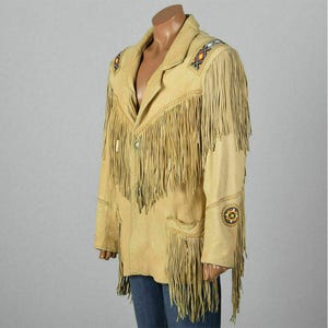 May include: Tan suede jacket with long fringe detailing on the chest, sleeves, and back. The jacket features decorative beadwork on the shoulders and sleeves. It has a classic collar and two front pockets.