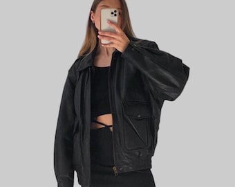 May include: Black leather bomber jacket with a collared neckline, two front pockets, and a zip closure. The jacket has a relaxed fit and is worn with a black top and skirt. The outfit is set against a plain gray background.
