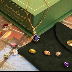 May include: A gold necklace with a square, deep blue gemstone pendant and a small diamond accent. The necklace is displayed with a green jewelry box and a dark green pouch. Scattered around are various colorful gemstones.