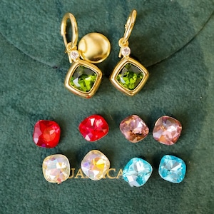 May include: Gold-tone earrings with square green gemstones and small clear accent stones. Additional loose gemstones in red, pink, and blue hues are also displayed. The earrings and gemstones are set against a dark green background.