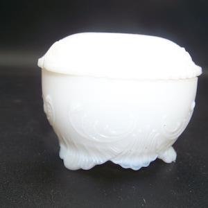 May include: White porcelain trinket box with a lid and ornate detailing. The box has a rounded shape and sits on three feet.