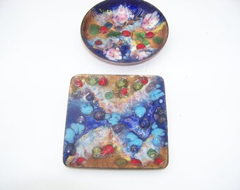 May include: Two decorative enamel pieces: a small, round dish and a square coaster. The dish features a blue background with floral and abstract designs in pink, red, and gold. The coaster has a similar design with blue, turquoise, red, and gold accents.