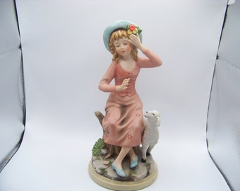 May include: A porcelain figurine of a girl wearing a light blue hat with flowers, a pink dress, and blue shoes. She is sitting on a tree stump with a white lamb beside her.