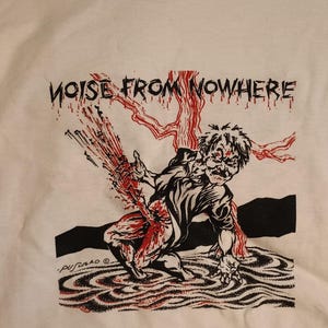 May include: White t-shirt with a graphic illustration featuring a figure with blood splatter and the text "NOISE FROM NOWHERE" in black. The design includes red and black elements, creating a striking visual.