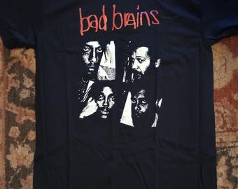 May include: Black t-shirt featuring the band name "bad brains" in red, above a white graphic of four faces. The shirt is made of a soft material and is likely a music-themed apparel item.