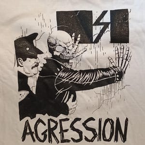 May include: White t-shirt featuring a black and white graphic illustration. The design includes a skull-like figure in a leather jacket, a police officer, and the word "AGGRESSION" in bold letters. The artwork has a punk rock aesthetic.