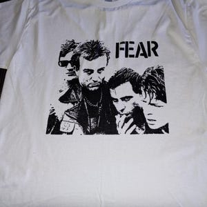 May include: White t-shirt featuring a black and white graphic of a group of people and the word "FEAR" in bold letters. The design appears to be a band or music-related theme, printed on a classic crew neck tee.