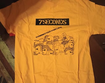 May include: Mustard yellow t-shirt with the text "7SECONDS" in a black rectangle above a graphic of several faces. Below the text is the phrase "COMMITTED FOR LIFE" in a curved font.