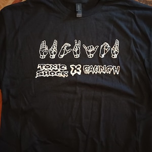 May include: Black t-shirt with white graphic text and hand signs. The text includes "TOXIC SHOCK" and "FAUNCH". The hand signs appear to spell out a word or phrase. The shirt is made of a soft material.