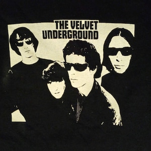 May include: Black t-shirt featuring a graphic of The Velvet Underground band members. The band members are depicted in a black and white style, wearing sunglasses. The band's name is printed in bold, white letters above the band members.