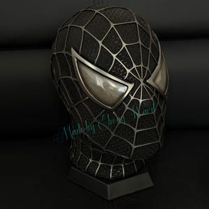 May include: A black Spider-Man mask with silver web detailing and large, reflective eye lenses. The mask is displayed on a black stand. The text "Made by Ebong Ward" is visible on the mask.