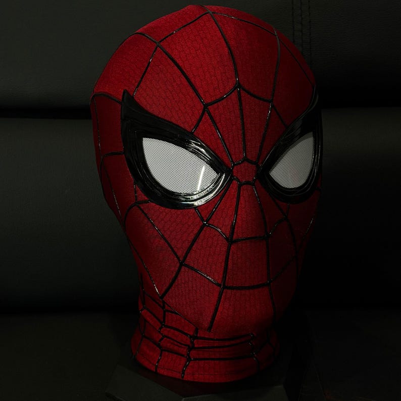 以下が含まれることがあります： 赤と黒のスパイダーマンマスク、クモの巣模様。 マスクには、黒で縁取られた白いメッシュの目があります。 マスクは、暗い背景に対して黒いスタンドに表示されています。