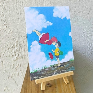 May include: A colorful illustration of two animated characters kissing in a blue sky with fluffy white clouds. One character is wearing a red dress and the other is wearing a yellow shirt. The artwork is displayed on a small wooden easel.