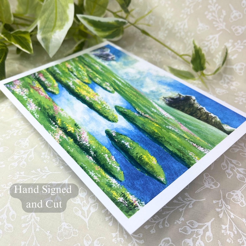 May include: A hand-signed and cut art print featuring a watercolor landscape. The artwork depicts green, elongated forms with pink accents against a blue sky and water. The print has a white border.